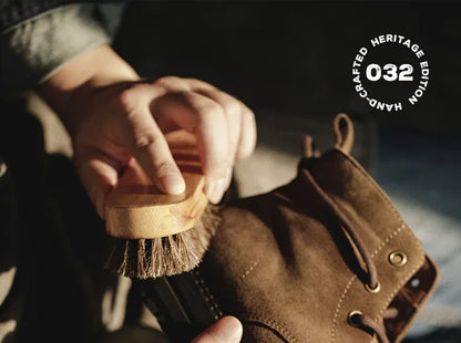 Person cleaning a brown leather boot with a brush, with 'Handcrafted Heritage Edition 032' logo in the corner.